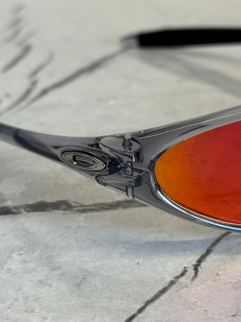 K&ouml;nnte beinhalten: Nahaufnahme einer Sonnenbrille mit transparentem grauem Rahmen und leuchtend roten Gl&auml;sern. Das Oakley-Logo ist auf dem B&uuml;gel zu sehen. Die Sonnenbrille befindet sich vor einem hellen, strukturierten Hintergrund.