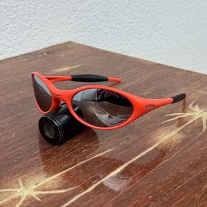 Pu&ograve; includere: Occhiali da sole rosso acceso con montatura curva e lenti scure. Gli occhiali presentano dettagli neri sulle aste e il logo Oakley &egrave; visibile. Sono appoggiati su un oggetto cilindrico nero.