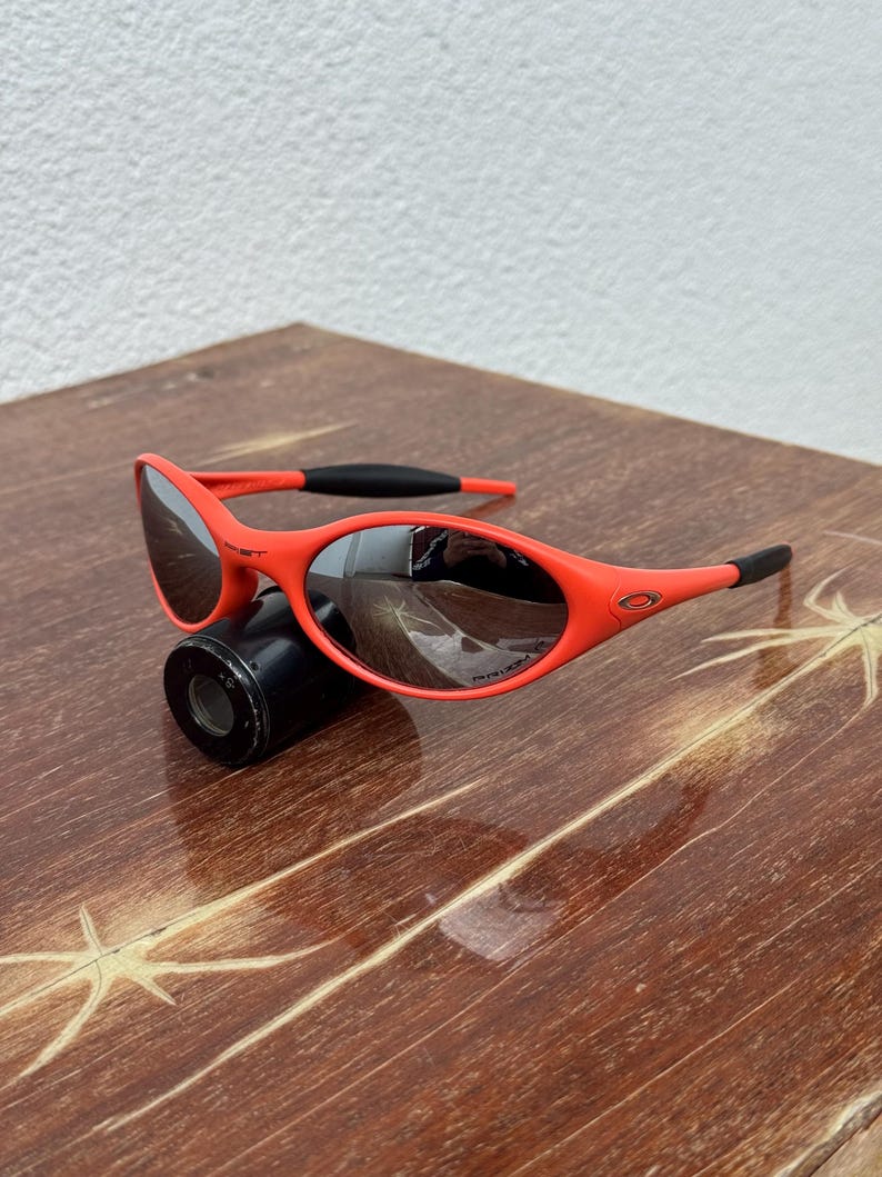 Pu&ograve; includere: Occhiali da sole rosso acceso con montatura curva e lenti scure. Gli occhiali presentano dettagli neri sulle aste e il logo Oakley &egrave; visibile. Sono appoggiati su un oggetto cilindrico nero.