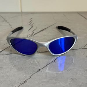 May include: Silver sunglasses with blue lenses. The sunglasses have a sleek, modern design with a curved frame and dark accents on the temples. The lenses reflect light, creating a vibrant blue hue.