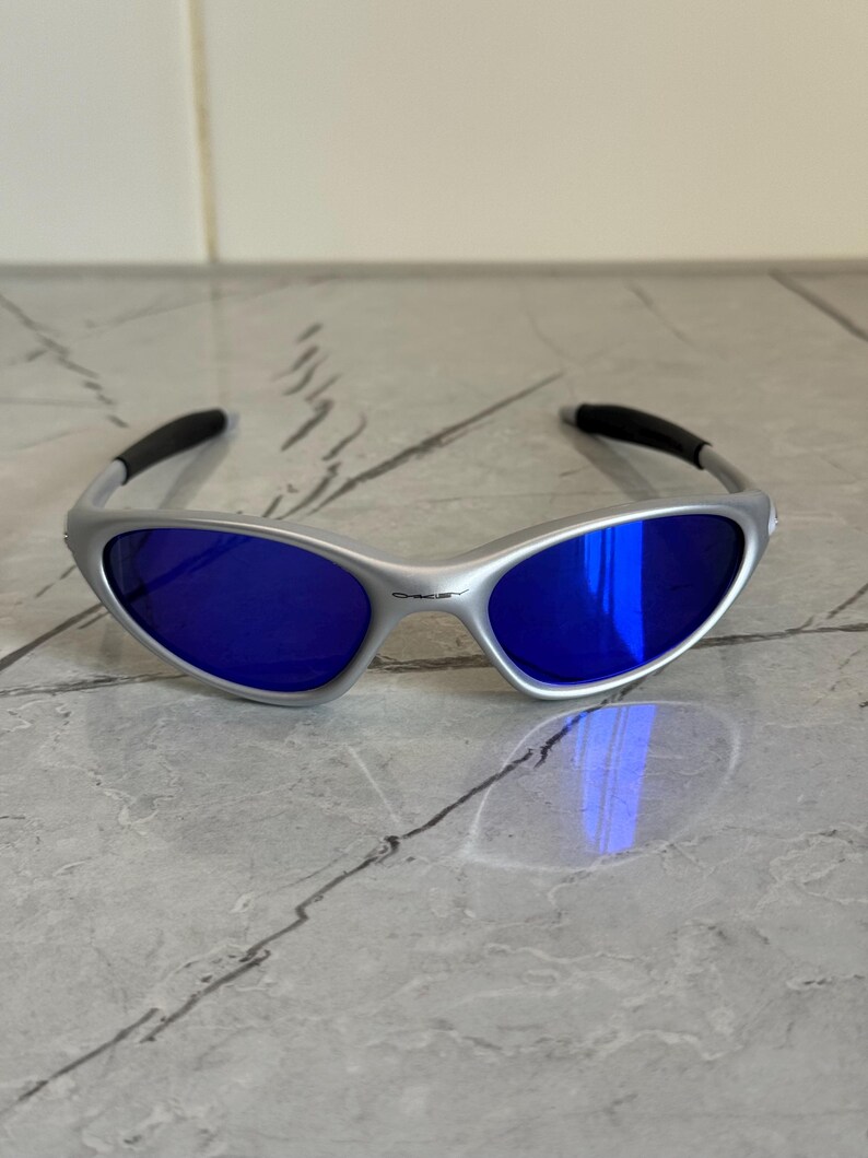 May include: Silver sunglasses with blue lenses. The sunglasses have a sleek, modern design with a curved frame and dark accents on the temples. The lenses reflect light, creating a vibrant blue hue.