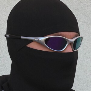 May include: Silver-framed sunglasses with purple and blue lenses. The sunglasses are worn over a black balaclava. The balaclava covers the head and neck, leaving only the eyes visible.