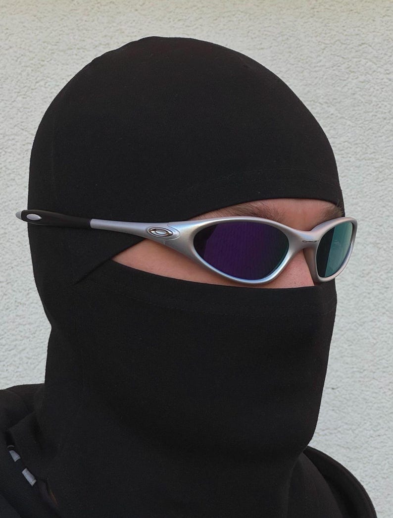 May include: Silver-framed sunglasses with purple and blue lenses. The sunglasses are worn over a black balaclava. The balaclava covers the head and neck, leaving only the eyes visible.