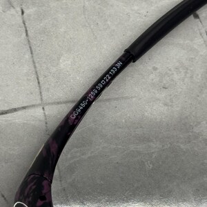 May include: Close-up of a black and purple patterned eyeglass frame. The frame has the text "DG9456-1259 59 22 133 3N" printed on the arm. The arm is black and the pattern is purple.