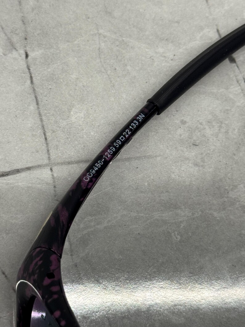 May include: Close-up of a black and purple patterned eyeglass frame. The frame has the text "DG9456-1259 59 22 133 3N" printed on the arm. The arm is black and the pattern is purple.