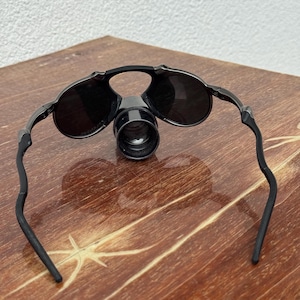 May include: Black magnifying glasses with round lenses and a central magnifying lens. The glasses have a sleek, modern design with curved temples. The lenses are dark, and the frame appears to be made of a durable material. The glasses are designed for close-up work.