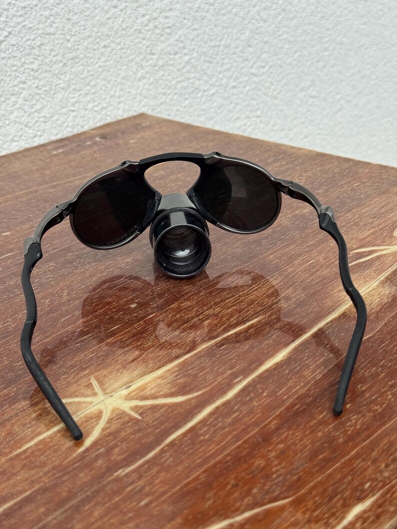 May include: Black magnifying glasses with round lenses and a central magnifying lens. The glasses have a sleek, modern design with curved temples. The lenses are dark, and the frame appears to be made of a durable material. The glasses are designed for close-up work.