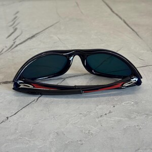 May include: Black sunglasses with dark lenses. The frame is black with red and silver accents on the arms. The sunglasses are lying on a gray surface.