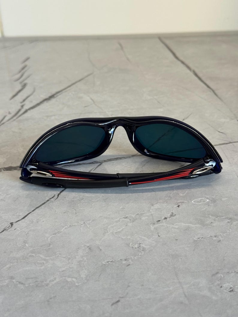 May include: Black sunglasses with dark lenses. The frame is black with red and silver accents on the arms. The sunglasses are lying on a gray surface.