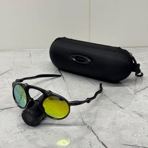 May include: Black Oakley sunglasses with yellow and green lenses, and a black case. The sunglasses have a unique design with a central cylindrical component and curved arms. The case is oval-shaped with a zipper closure.