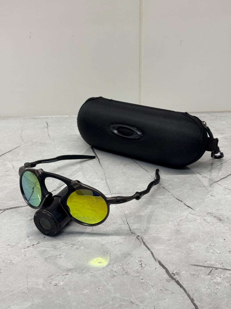 May include: Black Oakley sunglasses with yellow and green lenses, and a black case. The sunglasses have a unique design with a central cylindrical component and curved arms. The case is oval-shaped with a zipper closure.