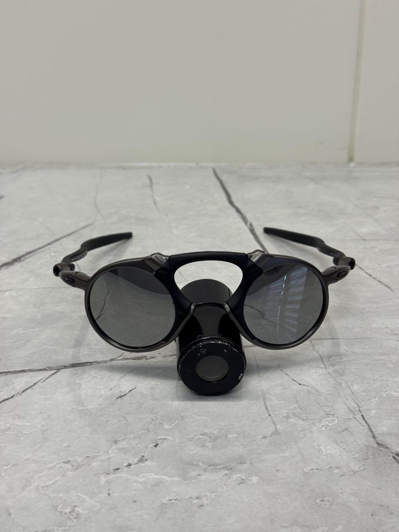May include: Black sunglasses with circular lenses and a distinctive bridge. The sunglasses have a matte finish and a futuristic design. The lenses are reflective, and the frame is made of a durable material.