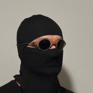 May include: A person wearing a black balaclava and sunglasses with round, dark lenses and a silver frame. The sunglasses have brown accents around the lenses. The person is wearing a black shirt.