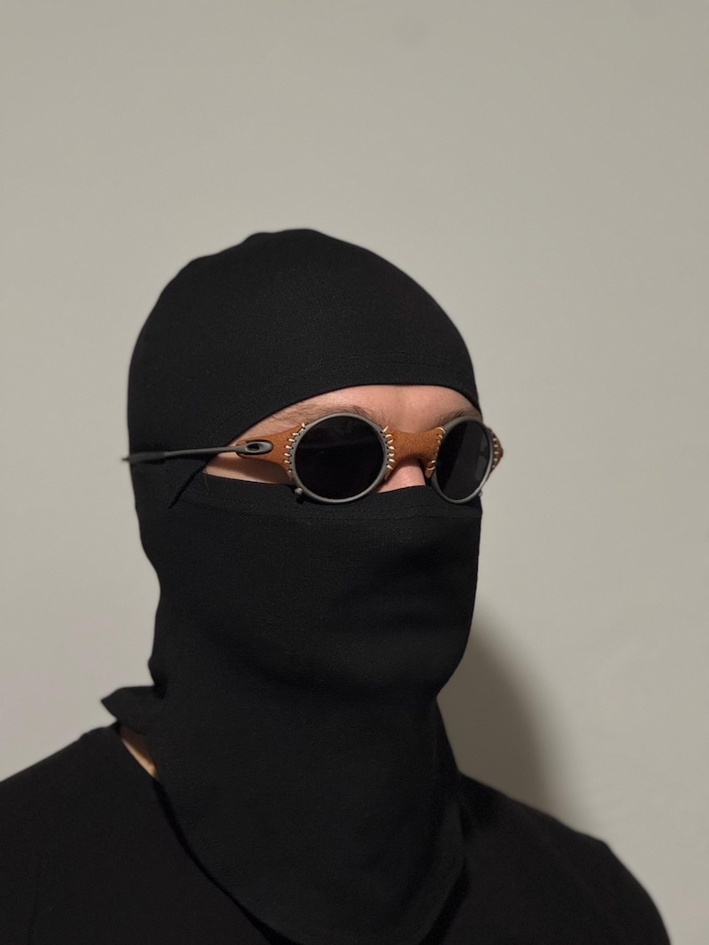 May include: A person wearing a black balaclava and sunglasses with round, dark lenses and a silver frame. The sunglasses have brown accents around the lenses. The person is wearing a black shirt.