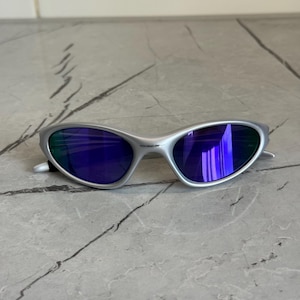 May include: Silver sunglasses with blue lenses. The sunglasses have a sleek, curved design and a metallic finish. The lenses reflect a vibrant blue hue, and the frame appears to be made of a lightweight metal.