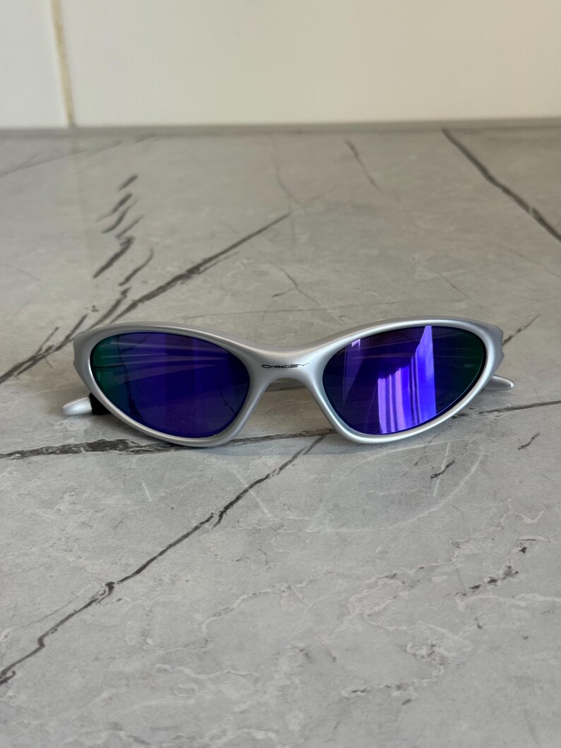 May include: Silver sunglasses with blue lenses. The sunglasses have a sleek, curved design and a metallic finish. The lenses reflect a vibrant blue hue, and the frame appears to be made of a lightweight metal.