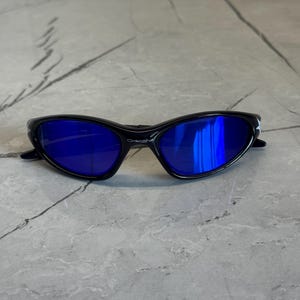 May include: Black sunglasses with blue lenses. The sunglasses have a sleek, curved design and the brand name "Oakley" is visible on the frame. The lenses reflect the surrounding environment.