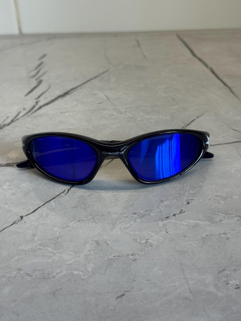 May include: Black sunglasses with blue lenses. The sunglasses have a sleek, curved design and the brand name "Oakley" is visible on the frame. The lenses reflect the surrounding environment.