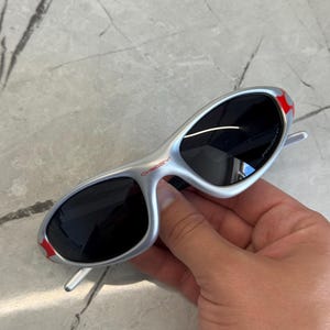 May include: Silver-framed sunglasses with black lenses. The sunglasses have red accents on the temples and the brand name "Oakley" is visible. The sunglasses are held in a hand.