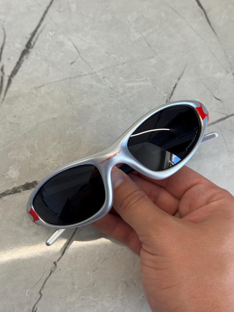May include: Silver-framed sunglasses with black lenses. The sunglasses have red accents on the temples and the brand name "Oakley" is visible. The sunglasses are held in a hand.