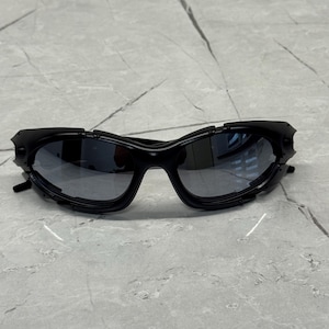 May include: Black sunglasses with a futuristic design. The sunglasses have a wraparound style with dark lenses and a textured frame. The frame features angular details and a sleek, modern aesthetic. The sunglasses are set against a neutral background.