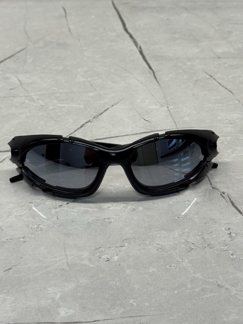 May include: Black sunglasses with a futuristic design. The sunglasses have a wraparound style with dark lenses and a textured frame. The frame features angular details and a sleek, modern aesthetic. The sunglasses are set against a neutral background.