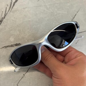 May include: Silver-framed sunglasses with dark lenses. The sunglasses have a sleek, curved design and the brand name "Oakley" is visible on the frame. The temples are white. The sunglasses are held by a hand.
