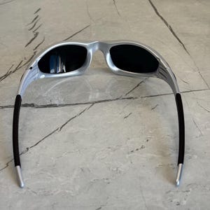 May include: Silver sunglasses with black lenses. The sunglasses have a sleek, modern design with a silver frame and black temples. The lenses are dark, providing protection from the sun. The sunglasses are designed for a comfortable fit.