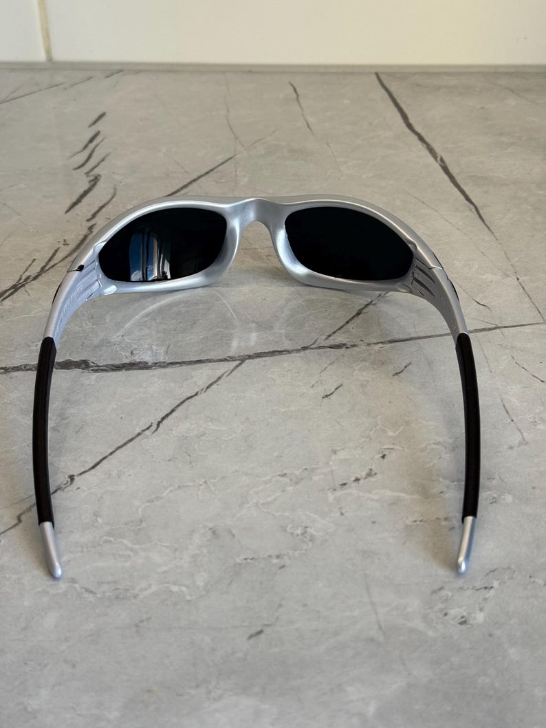 May include: Silver sunglasses with black lenses. The sunglasses have a sleek, modern design with a silver frame and black temples. The lenses are dark, providing protection from the sun. The sunglasses are designed for a comfortable fit.