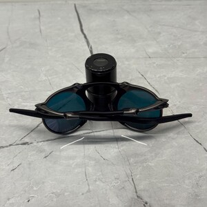 Pu&ograve; includere: Occhiali da sole neri con lenti blu. Gli occhiali hanno un design elegante e moderno con montatura e aste nere. Le lenti sono di un blu vibrante. Gli occhiali sono appoggiati su un oggetto cilindrico nero.