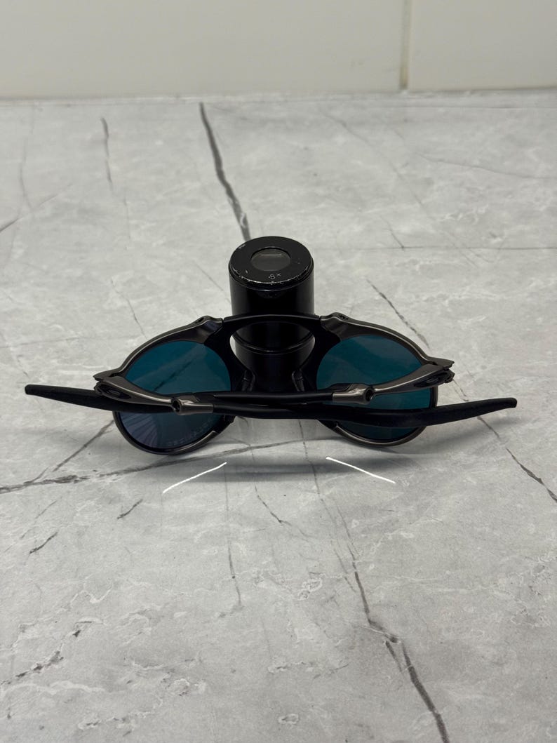 Pu&ograve; includere: Occhiali da sole neri con lenti blu. Gli occhiali hanno un design elegante e moderno con montatura e aste nere. Le lenti sono di un blu vibrante. Gli occhiali sono appoggiati su un oggetto cilindrico nero.