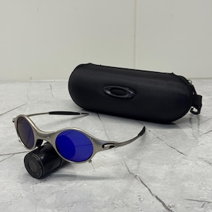 May include: Silver-framed sunglasses with blue lenses rest on a black cylindrical object. A black, oval-shaped case with a zipper and the Oakley logo sits behind the sunglasses. The sunglasses have a sporty, modern design.