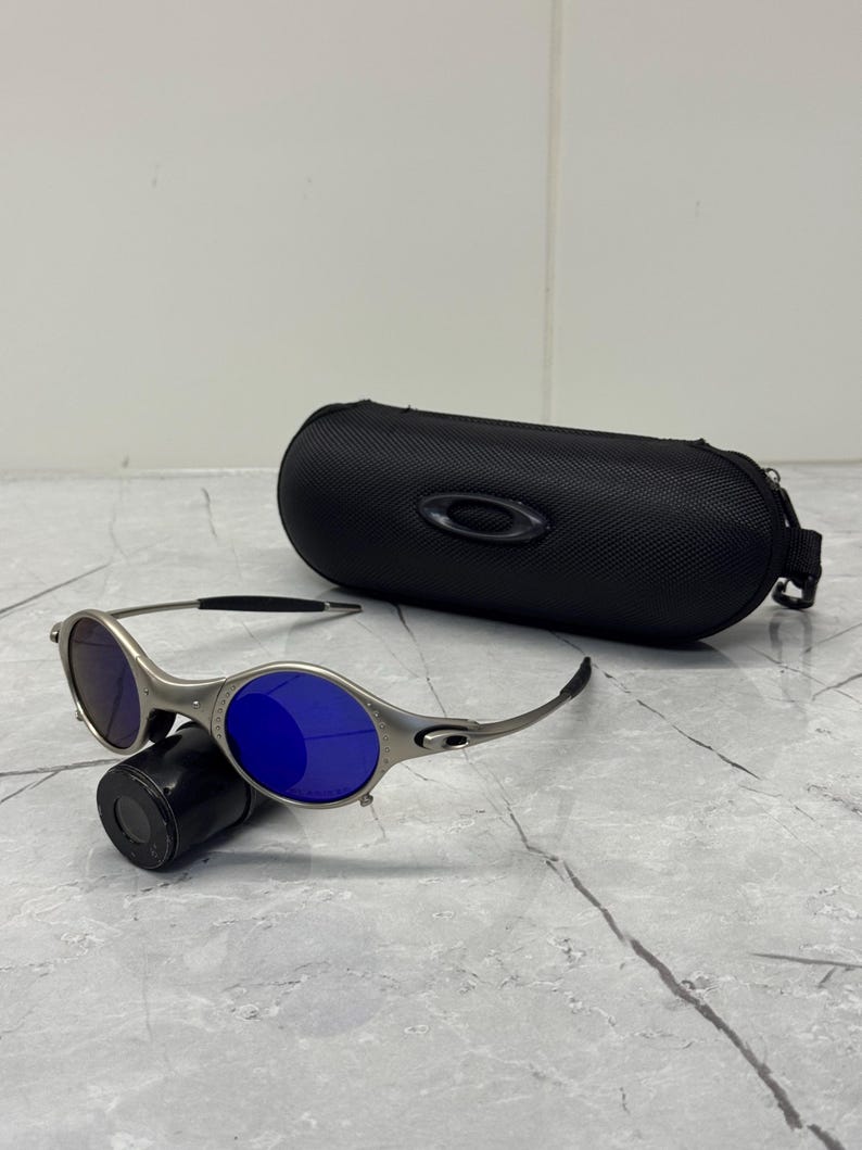 May include: Silver-framed sunglasses with blue lenses rest on a black cylindrical object. A black, oval-shaped case with a zipper and the Oakley logo sits behind the sunglasses. The sunglasses have a sporty, modern design.