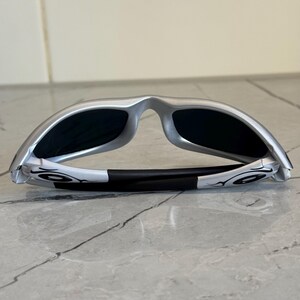 May include: Silver-framed sunglasses with black lenses. The sunglasses have a sleek, modern design with a black nose bridge and white accents on the temples. The lenses are dark, providing shade and protection from the sun.