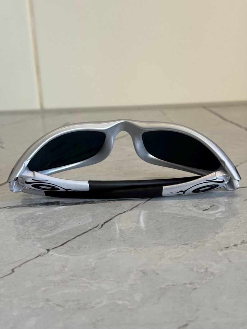 May include: Silver-framed sunglasses with black lenses. The sunglasses have a sleek, modern design with a black nose bridge and white accents on the temples. The lenses are dark, providing shade and protection from the sun.
