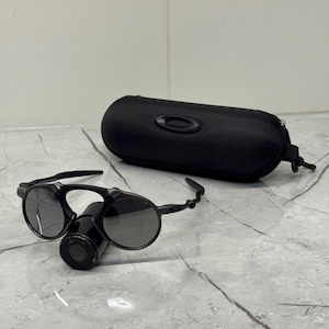 May include: Black sunglasses with circular lenses and a distinctive bridge design, next to a matching black case. The sunglasses have a sleek, modern look, suitable for outdoor activities. The case features the brand's logo.