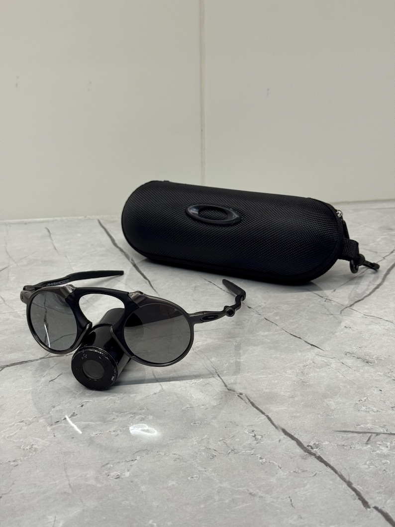 May include: Black sunglasses with circular lenses and a distinctive bridge design, next to a matching black case. The sunglasses have a sleek, modern look, suitable for outdoor activities. The case features the brand's logo.