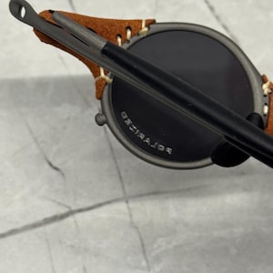 May include: Close-up of round sunglasses with dark lenses and a matte black frame. The temples are attached to a brown suede leather piece. The text "POLARIZED" is visible on the lens.
