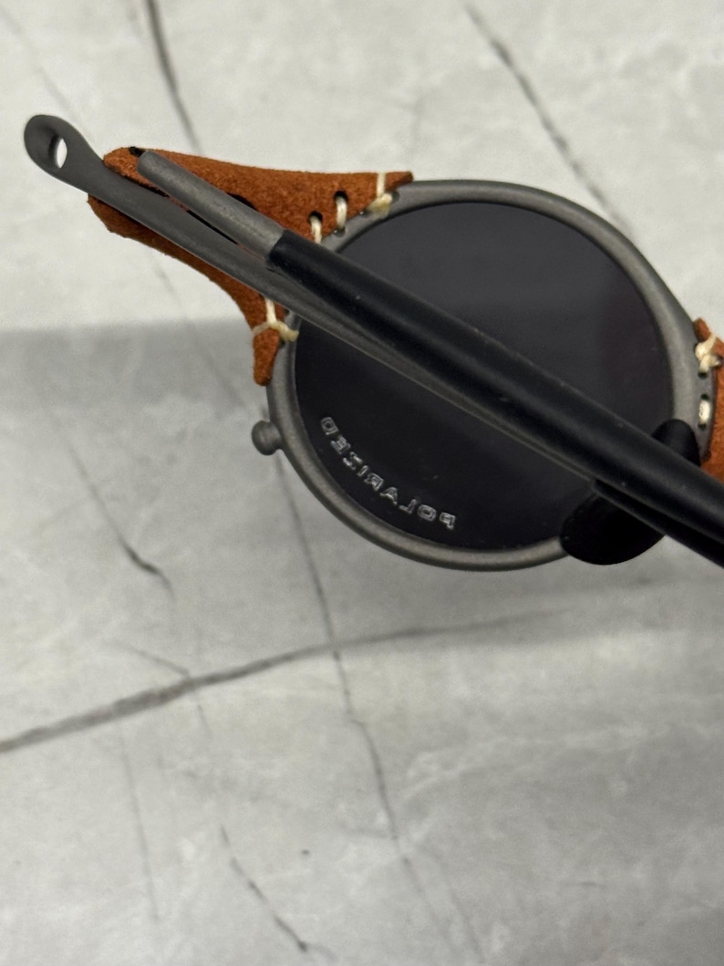 May include: Close-up of round sunglasses with dark lenses and a matte black frame. The temples are attached to a brown suede leather piece. The text "POLARIZED" is visible on the lens.