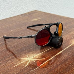May include: Black and silver-toned sunglasses with red and orange lenses. The sunglasses have a unique design with a circular lens and a cylindrical attachment. The frame is a dark metallic color, and the lenses are a vibrant red and orange.