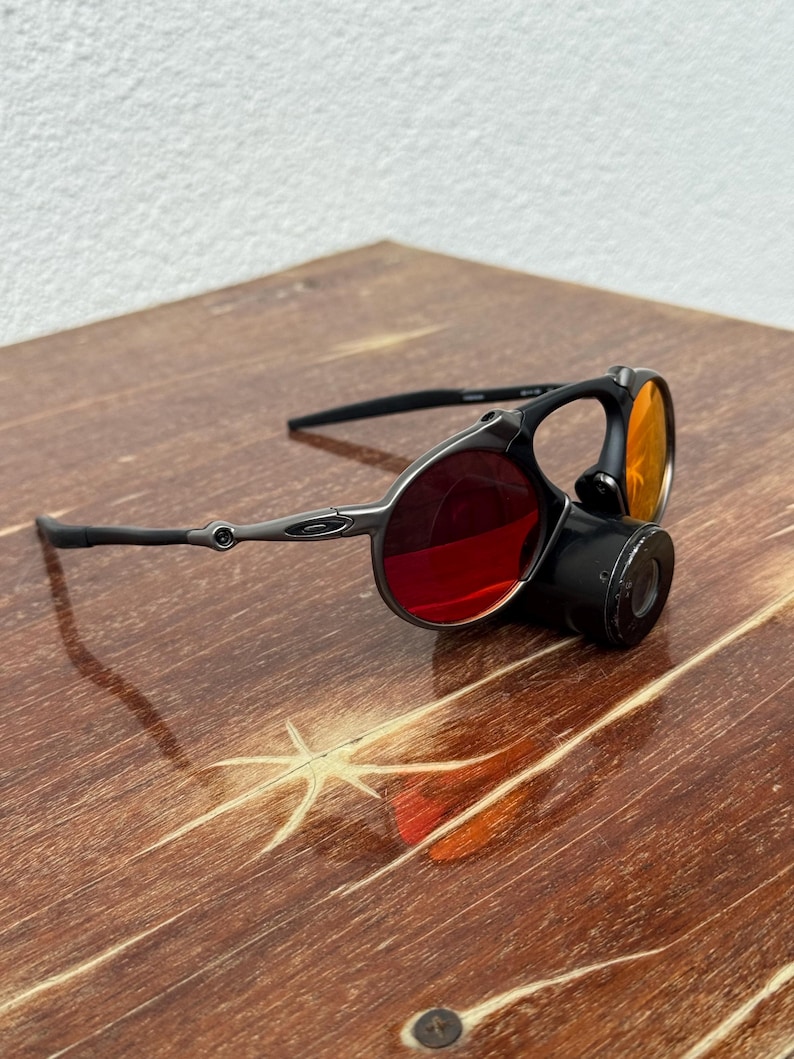 May include: Black and silver-toned sunglasses with red and orange lenses. The sunglasses have a unique design with a circular lens and a cylindrical attachment. The frame is a dark metallic color, and the lenses are a vibrant red and orange.