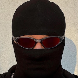 K&ouml;nnte beinhalten: Eine Person tr&auml;gt eine schwarze Sturmhaube und eine Brille mit transparentem Rahmen und roten Gl&auml;sern. Die Sturmhaube bedeckt Kopf und Hals, mit &Ouml;ffnungen f&uuml;r Augen und Mund. Die Sonnenbrille ist &uuml;ber den Augen positioniert.
