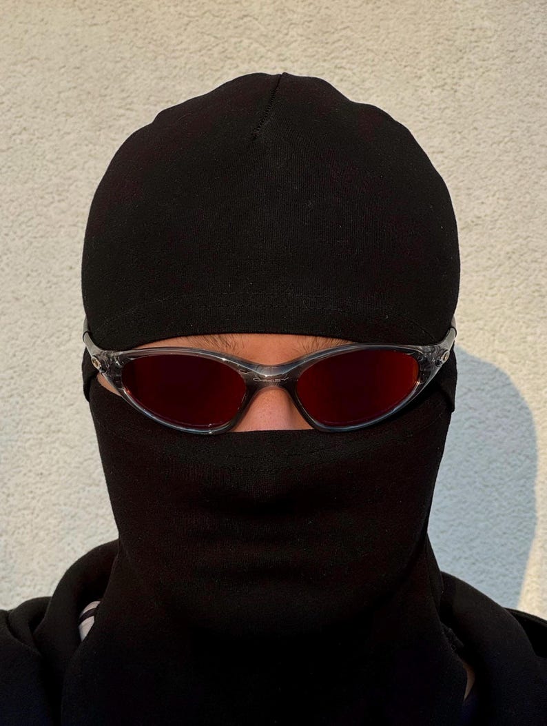 K&ouml;nnte beinhalten: Eine Person tr&auml;gt eine schwarze Sturmhaube und eine Brille mit transparentem Rahmen und roten Gl&auml;sern. Die Sturmhaube bedeckt Kopf und Hals, mit &Ouml;ffnungen f&uuml;r Augen und Mund. Die Sonnenbrille ist &uuml;ber den Augen positioniert.
