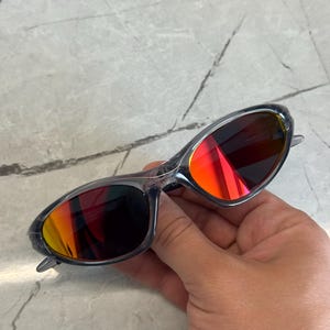 K&ouml;nnte beinhalten: Sonnenbrille mit transparentem Rahmen und leuchtend roten und orangen Gl&auml;sern. Die Sonnenbrille hat ein schlankes, modernes Design. Die Gl&auml;ser reflektieren einen Farbverlauf. Der Rahmen ist transparent grau.