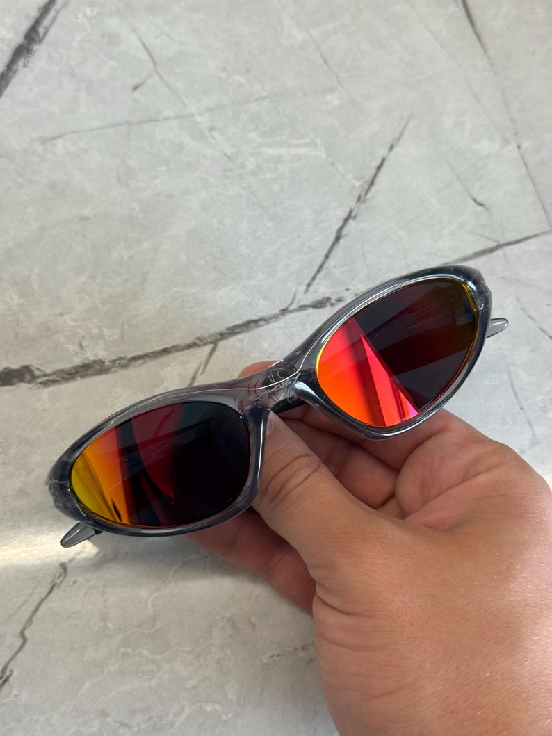 K&ouml;nnte beinhalten: Sonnenbrille mit transparentem Rahmen und leuchtend roten und orangen Gl&auml;sern. Die Sonnenbrille hat ein schlankes, modernes Design. Die Gl&auml;ser reflektieren einen Farbverlauf. Der Rahmen ist transparent grau.