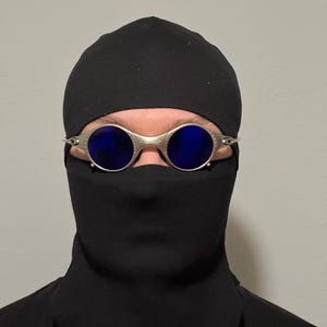May include: Silver-framed sunglasses with blue lenses are worn with a black head covering and face mask. The sunglasses have a round shape and a metallic finish. The head covering and face mask are made of a dark fabric.