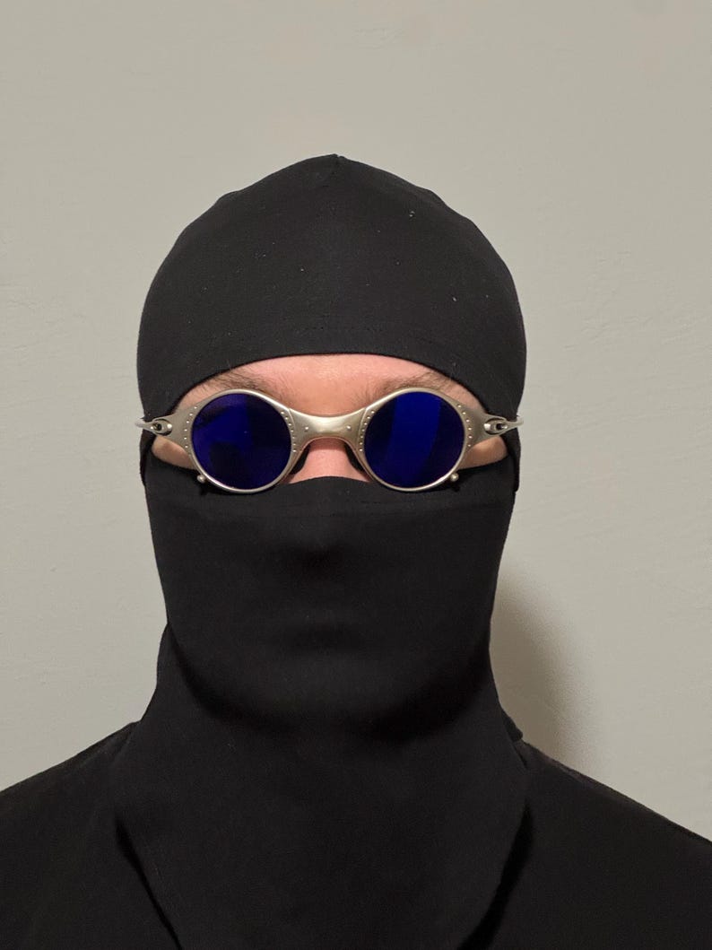 May include: Silver-framed sunglasses with blue lenses are worn with a black head covering and face mask. The sunglasses have a round shape and a metallic finish. The head covering and face mask are made of a dark fabric.