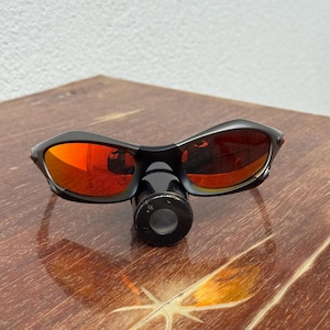 Op de afbeelding: Zwarte zonnebril met rood getinte glazen ligt op een houten oppervlak. De zonnebril heeft een gebogen ontwerp. Een klein, zwart, cirkelvormig object is aan de bril bevestigd. De glazen reflecteren de omgeving.