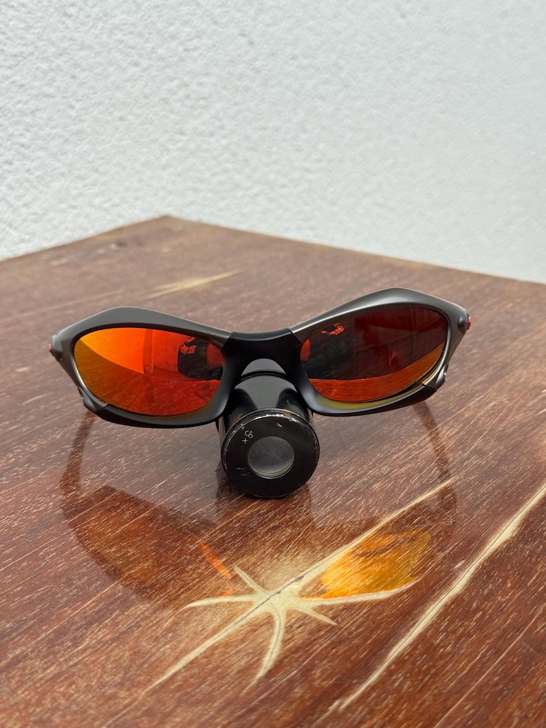 Op de afbeelding: Zwarte zonnebril met rood getinte glazen ligt op een houten oppervlak. De zonnebril heeft een gebogen ontwerp. Een klein, zwart, cirkelvormig object is aan de bril bevestigd. De glazen reflecteren de omgeving.