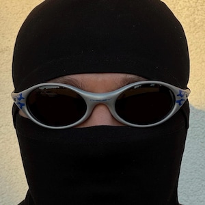 Puede incluir: Gafas de sol con montura plateada, lentes oscuras y detalles azules. Las gafas se llevan con un pasamonta&ntilde;as negro. La imagen es un primer plano que se centra en las gafas y el pasamonta&ntilde;as.